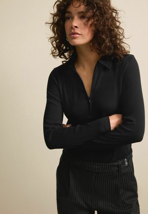Zwarte zip-up top met lange mouwen, geribbelde textuur en een kraaghalslijn. Gekozen in combinatie met zwart gestreepte broek met knoopaccenten.