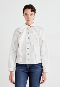Blusa blanca con volantes de encaje, lazo en el cuello, botones negros y detalles de ojal. Manga larga con puños fruncidos.