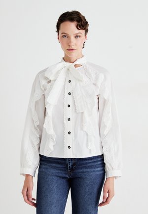 Hvit bluse med volangspiss trim, sløyfebånd i halsen, svarte knapper og blonderdetaljer. Lange ermer med samlet mansjett.
