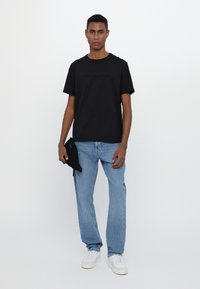 Camiseta de algodón negra con un logo en el frente, combinada con jeans de mezclilla azules claros y zapatillas blancas, sosteniendo un pequeño accesorio negro.