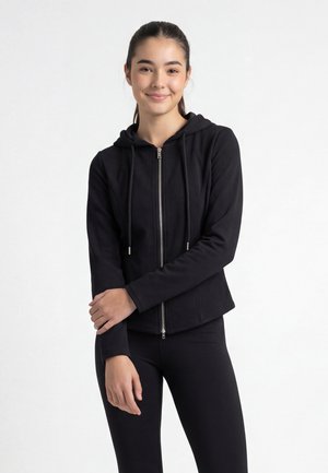 Jeune femme aux cheveux foncés attachés, portant un sweat à capuche noir zippé et un legging noir, debout devant un fond blanc uni.