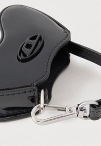 Príslušenstvo v tvare srdca z čiernej lakovanej kože s centrálnou prackou a striebornou kovovou sponou pripevnenou k čiernemu remienku.