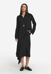 Svart skjorta och asymmetrisk kjol i mjukt tyg, med en knäppning fram och en elegant drapering. Tillsammans med svarta slipskor.