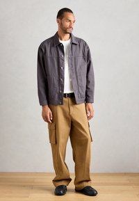 Man wearing a gray buttoned jacket, white shirt, tan cargo pants, black belt, and black shoes standing on a wooden floor against a plain wall.