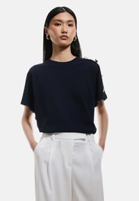 Marinblå kortärmad topp i slätt tyg, med rund halsringning och dekorativa knappar på axeln. Kombinerad med vita byxor.