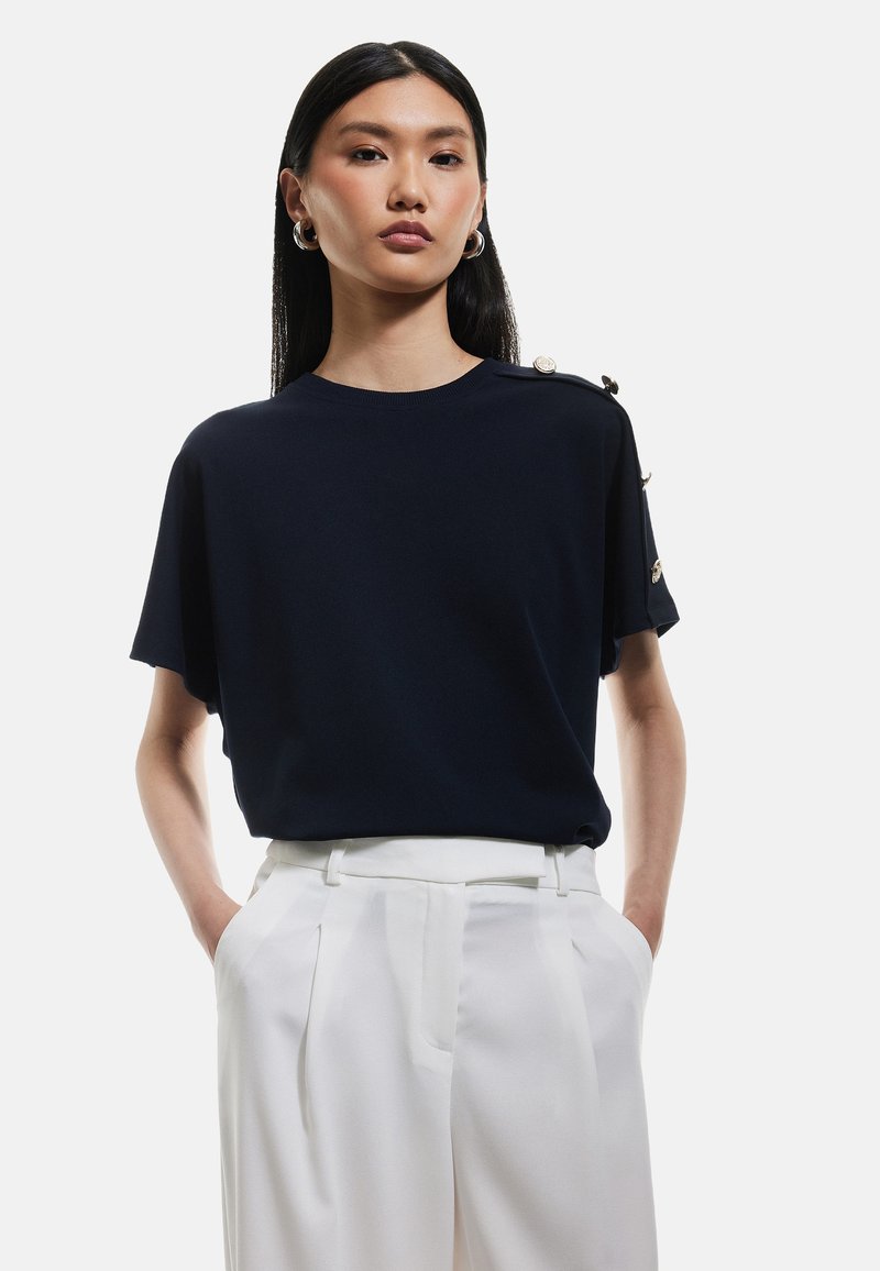 Marinblå kortärmad topp i slätt tyg, med rund halsringning och dekorativa knappar på axeln. Kombinerad med vita byxor.