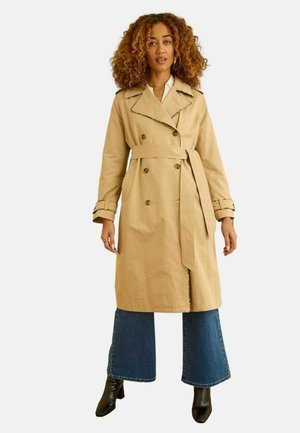 Frau mit lockigem Haar, die einen beigen Trenchcoat mit Gürtel, blaue Jeans mit weitem Bein und schwarze Stiefeletten mit Absatz trägt und vor weißem Hintergrund steht.