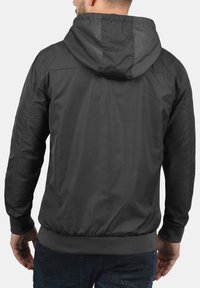 Mörkgrå hujacka med en slät, glänsande yta, utrustad med elastiska muddar och fåll, samt en enkel och funktionell design.