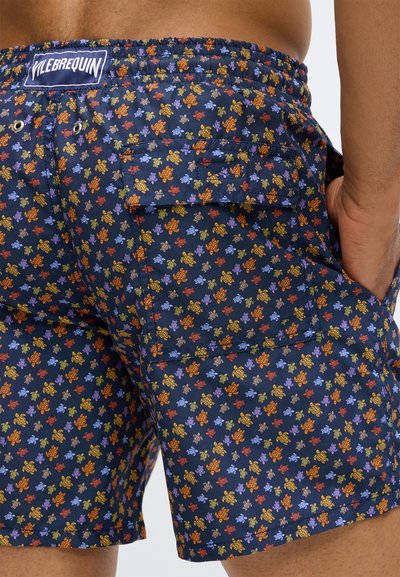 Prim-plan al șorturilor de baie de culoare bleumarin cu un model colorat de țestoase, buzunar posterior cu clapa și eticheta brandului pe talie, mână în buzunarul lateral.