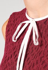 Haut en dentelle bourgogne avec un ruban blanc noué au cou. La texture est intricée, présentant un délicat motif floral tout au long.