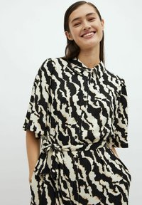 Černo-bílá vzorovaná košilová šaty s krátkými rukávy, předním zapínáním na knoflíky a uvázaným pasem. Tkanina se zdá být lehká a texturovaná.