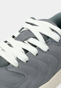 Baskets en suède gris avec des lacets blancs. Elles présentent une surface texturée et un col rembourré, avec une semelle en caoutchouc gris clair.