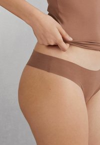 Honingkleurige naadloze strings gemaakt van rekbare stof. Het heeft een laag gesneden ontwerp met een gladde tailleband en minimale bedekking.
