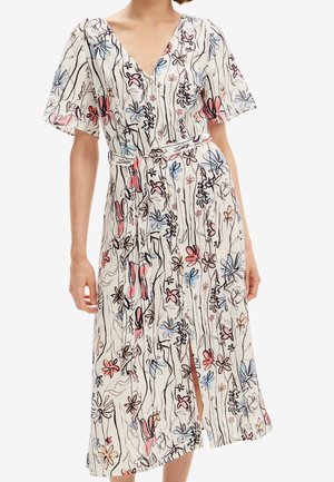 Žena v bílé midi šatech s krátkými rukávy, výstřihem do V, zavazováním v pase a barevným květinovým a čárovým vzorem.