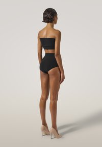 Svart tvådelad bikini med strapless topp och högmidjade bottoms. Slät textur, minimalistisk design och nakna högklackade skor. Neutral bakgrund.