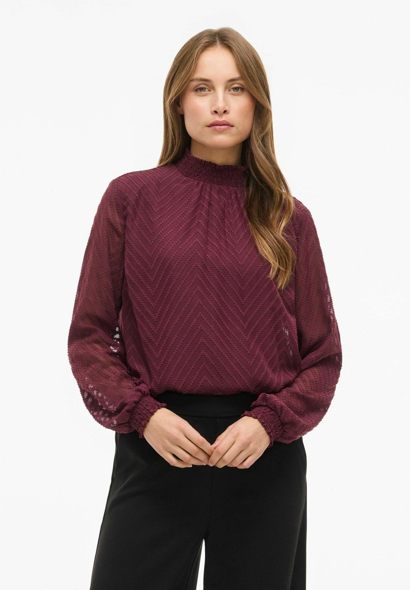 Femme aux longs cheveux portant un chemisier bordeaux texturé à manches longues avec des poignets volantés et un pantalon noir, debout devant un fond blanc.
