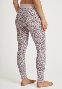 Vrouw in hoog getailleerde, roze leggings met een klein zwart bladerenpatroon, staand op blote voeten tegen een effen lichte achtergrond.