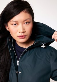 Veste imperméable vert foncé avec un col haut, fermeture éclair argentée et boutons-pressions, dotée d'une texture lisse et d'un cordon de serrage ajustable.