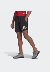 Shorts sportivi neri con vita elastica, con un grande logo Adidas bianco sulla gamba sinistra. Scarpe da ginnastica grigie con dettagli arancioni.