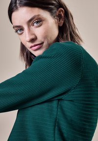 Pull à col rond en teal avec des côtes, tissu texturé, coutures angulaires et silhouette ajustée. Gros plan sur le détail de la manche.