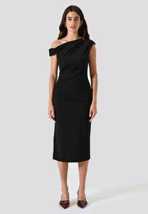 Robe noire à épaules dénudées en tissu lisse, design ajusté, longueur mi-mollet, avec des détails texturés et une fente sur le côté. Associée à des talons marron.