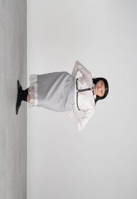 Eine Frau steht mit den Händen in den Hüften, trägt eine weiße Bluse, einen grauen Rock mit Spitzenbesatz, schwarze Socken und schwarze Schuhe vor einem schlichten grauen Hintergrund.