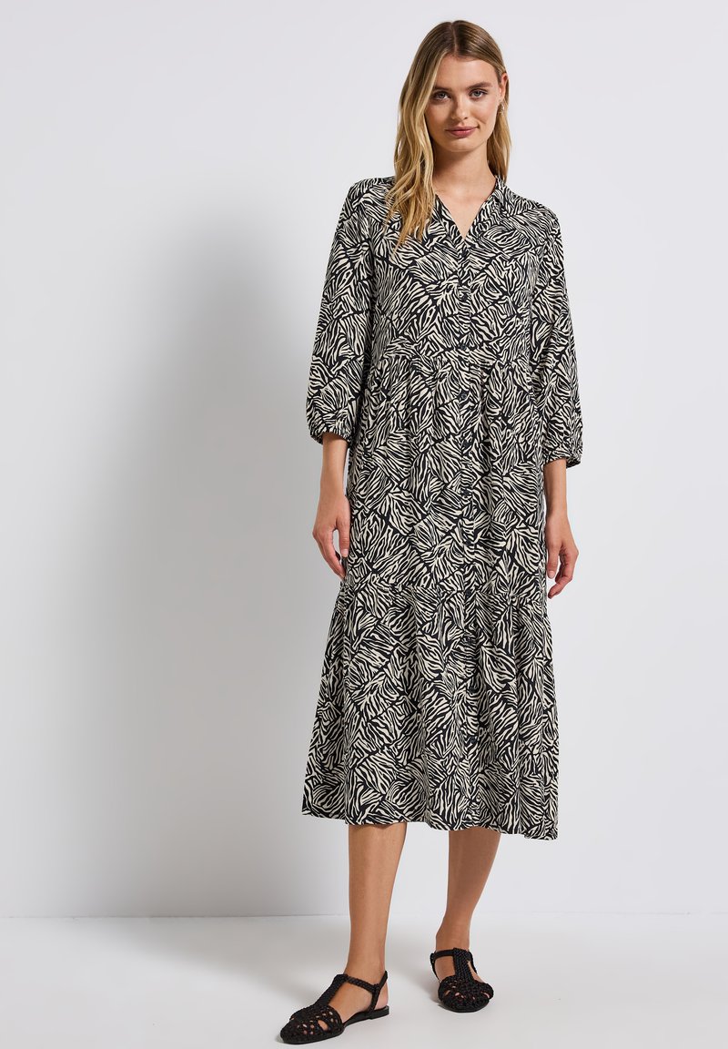 Abito maxi in bianco e nero con un motivo a foglie, maniche lunghe a sbuffo, scollatura a V e design della gonna a strati. Indossato con sandali neri.
