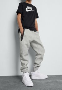 Svart Nike-t-shirt med vit logotyp, grå mjukisbyxor med blixtlåsficka och vita Nike högklackade sneakers, alla stående på en grå yta.