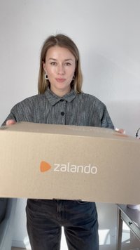 Caixa de cartão castanho com o logo da Zalando, segurada por uma pessoa com uma camisa cinza texturizada. A caixa é retangular e parece estar selada.