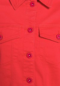 Chaqueta de mezclilla roja con botones rojos, dos bolsillos en el pecho con solapas y un cuello puntiagudo visible.