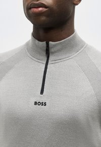 Gros plan sur une personne portant un pull zippé gris clair avec un col côtelé et le logo "BOSS" sur la poitrine.