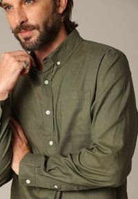 Olijfgroen button-up shirt van zachte stof, voorzien van een borstzak, lange mouwen, knoopmanchetten en witte knopen.