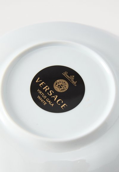 Balta keramikas šķīvis ar spīdīgu pārklājumu. Apaļa forma un melna etiķete ar uzrakstiem "Versace", "Virtus Gala" un logotipu.