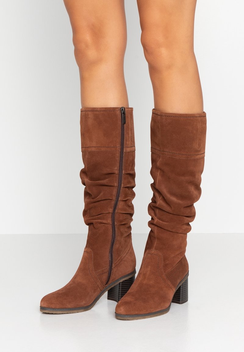 Jambes portant des bottes montantes en suède marron avec talons carrés et fermetures éclair latérales, debout sur un sol blanc contre un fond uni.