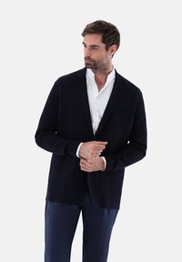 Marineblaue Strickjacke aus weichem Strick mit Schalkragen, offenem Vorderteil und zwei Fronttaschen, über einem weißen Hemd getragen.