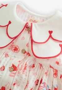 Balta kleita ar sarkanu viļņotu apkakli un ziedu rakstiem sarkanā un zilā krāsā. Pogas aizdare un krokota tekstūra piešķir detaļas.