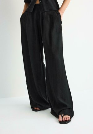 Schwarze Hosen mit weitem Bein und vorderen Falten, getragen mit passendem schwarzen, knöpfbarem Oberteil und schwarzen Sandalen mit quadratischer Spitze. Hände in den Taschen.