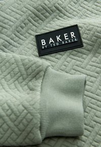 Gai zaļš teksturēts džemperis ar pārliktu rakstu un melnu birku "BAKER by TED BAKER." Redzams ribots aproces detaļas.