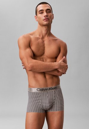 Mężczyzna bez koszuli z krótkimi włosami, ubrany w wzorzyste bokserki Calvin Klein, stojący z założonymi ramionami na tle jednolitego szarego tła.