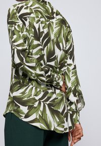 Chemise à manches longues en tissu blanc avec un motif de feuilles vertes. L'arrière présente un ourlet incurvé et des accents de boutons sur les poignets.