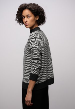Schwarz-weiß gemusterter Pullover mit Chevron-Muster, Rundhalsausschnitt, strukturierter Strick, gerippte Bündchen und Saum, lockere Passform. Modell posiert seitlich.