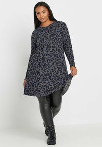 Langärmliges graues Kleid mit schwarzem und blauem Leopardenmuster, gekurvtem Saum und strukturierter Stoffqualität, kombiniert mit schwarzen Leggings und Ankle Boots.