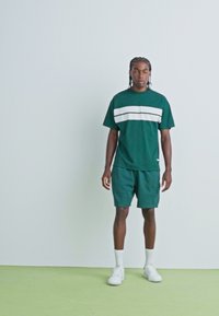 Zöld, két részes sportos szett, amely tartalmaz egy crew neck pólót fehér csíkokkal és hozzá illő sortot, sima anyagból készült. Fehér tornacipőt visel.