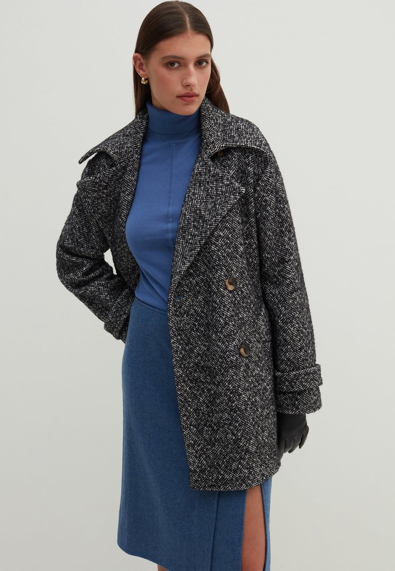 Cappotto houndstooth in bianco e nero con ampi revers, bottoni doppi e tessuto scuro; indossato sopra un dolcevita blu e una gonna.