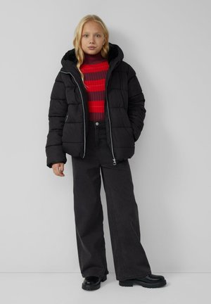 Doudoune noire avec capuche, présentant une texture matelassée et une fermeture éclair. Associée à un col roulé rayé rouge et bordeaux et à un jean large noir.