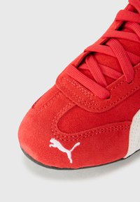 Baskets en daim rouge avec lacets, logo Puma blanc, forme courbée et surface texturée. Semelle noire visible au fond.