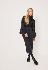 Crna traper jakna preko crne dolčevite, uparena s crnim hlačama viskog struka i crnim tenisicama. Minimalistički stil, neutralne boje.