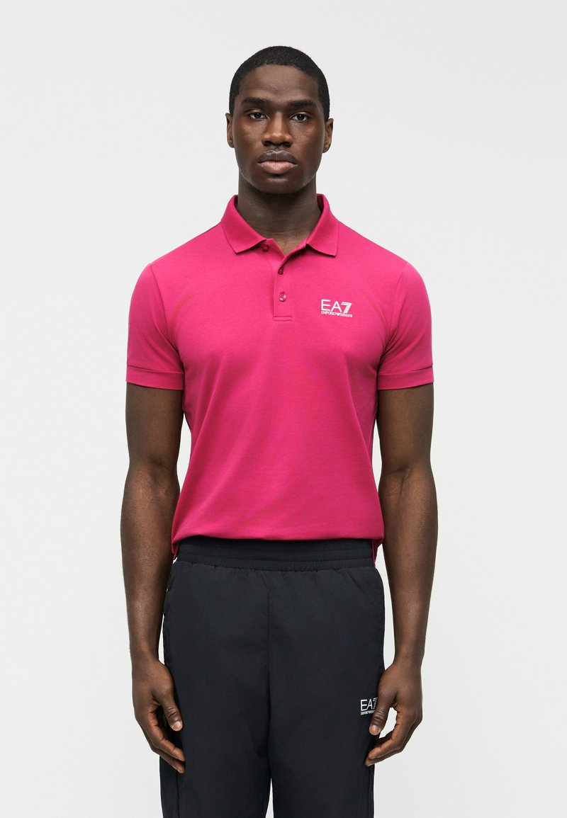 Άνδρας που φοράει εφαρμοστή φωτεινή ροζ πόλο μπλούζα EA7 και μαύρο παντελόνι EA7, στέκεται μπροστά σε απλό φωτεινό φόντο.
