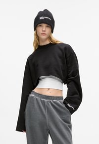 Črn skrajšan sweatshirt z dolgimi rokavi, v kombinaciji s sivimi žametnimi hlačami. Klobuk ima belo besedilo, kar dodaja kontrast oblačilu.