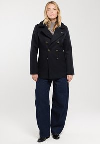 Abrigo de marina cruzado en color azul marino con botones dorados, solapas anchas y un corte estructurado, combinado con pantalones vaqueros de denim azul sueltos y botas negras.